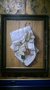 A bundle of documents suspended from a nail on a plain wooden board in a painted (trompe l'oeil) frame